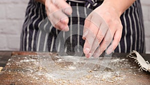 Woman in a blue apron claps her hands and white wheat flour flies in different directions over a wooden table