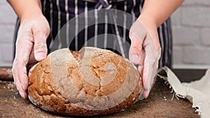 A woman in a blue apron holds baked round rye flour bread in her hands over the table
