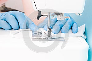 a woman in blue disposable medical gloves sews on a sewing machine a protective face mask made of cotton gray fabric