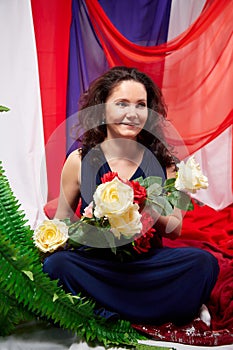 Woman in blue dress with long brunette curly hair and roses in hands indoors with red and blue background. Model posing