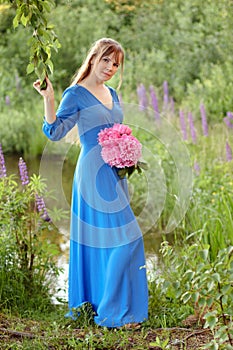 Woman in blue dress in purple lupines field. Meadow of violet flowers in the summer. Girl with long hair holding a lupine bouquet