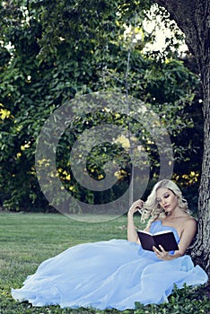 Woman in blue dress reading a book leaning on tree
