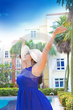 Woman in blue dress and white hat with arms wide apart happy by the pool