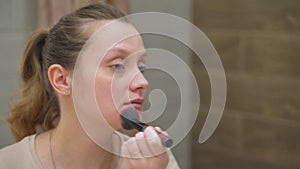 Woman with blue eyes is powdering the skin on face with fluffy brush, standing by large mirror in the bathroom. Her wavy