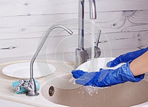 A woman in blue gloves washes a white plate under a stream of water over the sink