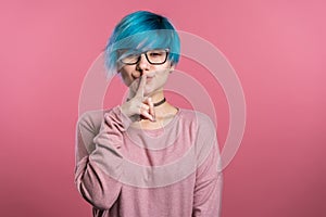 Woman with blue hair holding finger on her lips over pink background. Gesture of shhh, secret, silence. Close up.