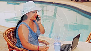 A woman in a blue hat sits at a table by the pool and works with a laptop remotely