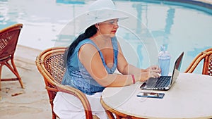 A woman in a blue hat sits at a table by the pool and works with a laptop remotely