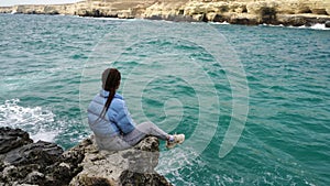 A woman in a blue jacket sits on a rock above a cliff above the sea, looking at the stormy ocean. Girl traveler rests