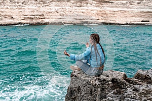 A woman in a blue jacket sits on a rock above a cliff above the sea, looking at the stormy ocean. Girl traveler rests