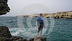 A woman in a blue jacket walks on a cliff above a cliff overlooking the sea, looking at the stormy ocean. Girl traveler