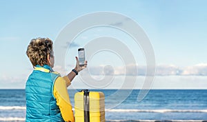 Woman in blue jacket with a yellow suitcase and headphones on the seaside on hot sunny day arrived in a tourist town. Travel