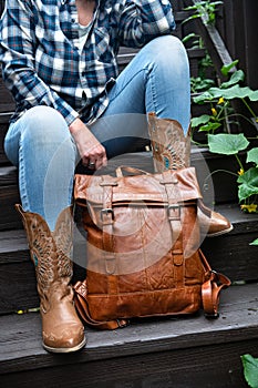 woman in blue jeans and cowboy boots sits on the steps and holds a leather backpack. Close-up
