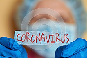 Woman in blue medical gloves, medical mask and safety cap is holding inscription Coronavirus on beige background. Closeup