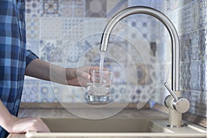 Woman in a blue plaid shirt pours water into the glass of the faucet in the kitchen