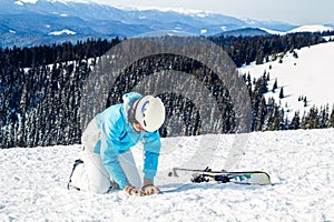 Woman in blue suit, helmet and glasses sits on the snow near the skis on top of the mountain