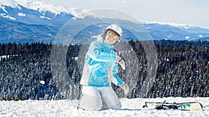 Woman in blue suit, helmet and glasses sits on the snow near the skis on top of the mountain