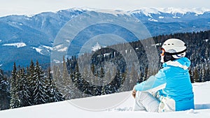 Woman in blue suit, helmet and glasses sits on the snow near the skis on top of the mountain