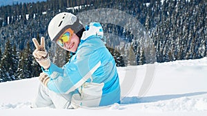 Woman in blue suit, helmet and glasses sits on the snow near the skis on top of the mountain