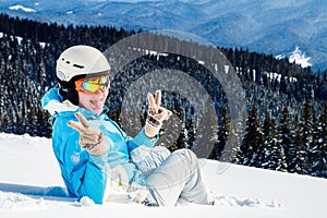 Woman in blue suit, helmet and glasses sits on the snow near the skis on top of the mountain