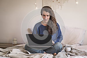 Woman in blue sweater casually working on laptop whilst sat on bed