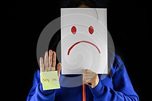 A woman in a blue sweater covering and hiding her face with a white cardboard with a sad face drawing sign representing depressio