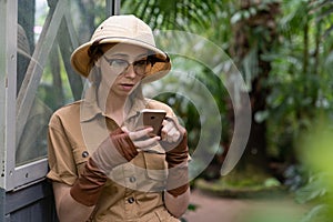 Woman botanist dressed in safari style in greenhouse. Naturalist in khaki clothes, leaver gloves resting and reading a book photo