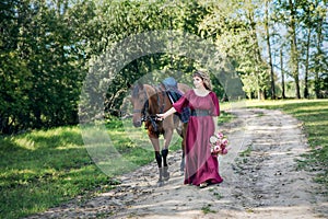 A woman with a bouquet of flowers dressed in a long burgundy dress with sleeves leads a brown horse