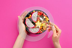 Woman with bowl of delicious exotic fruit salad on pink background, top view