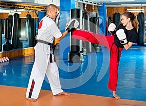 Woman in boxing gloves practicing kick with trainer holding focus mitts