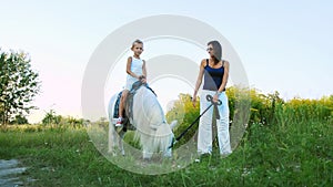 A woman and a boy are walking around the field, son is riding a pony, mother is holding a pony for a bridle. Cheerful