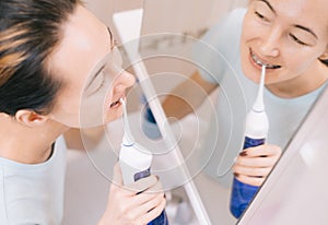 Woman with braces on her teeth brushing her teeth with by using a irrigate, before mirror, top view.