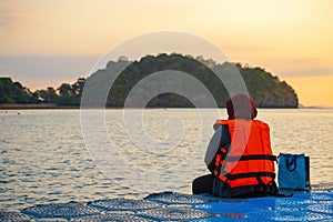 A woman in a bright orange life jacket sits on a pontoon pier early in the morning during sunrise. Dangling legs in the water and