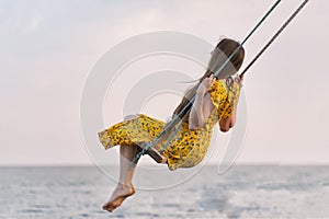 Woman in bright yellow dress rides on swing against the sea background. Alone with thoughts. Loneliness