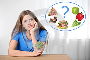 Woman with broccoli thinking about what to chose - healthy and unhealthy food at table indoors