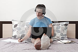 A woman with a broken leg works from home. A woman with headphones is sitting on the bed and holding a laptop on her lap