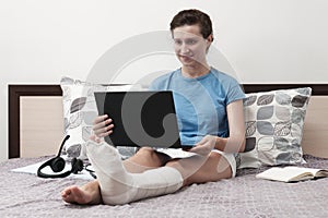 A woman with a broken leg works from home. A woman sitting on the bed and holding a laptop on her lap