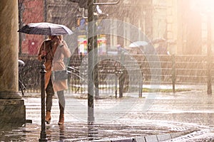 Woman in a brown coat under an umbrella crosses a busy street in heavy rain. Heavy precipitation in the city. Flooding of city