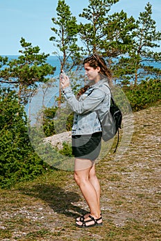 Woman with brown curly hair hiking and taking pictures with phone of the seaside by the Panga cliff forest in Saaremaa, Estonia