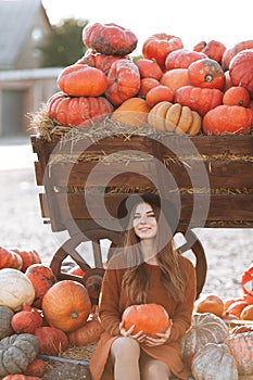 Woman in brown dress and sweater holding orange pumpkin and sitting near wooden wagon with plenty pumpkins on farmers market. Cozy