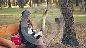 A woman with brown hair in a hat in red boots and a red bag on a park bench in autumn reads