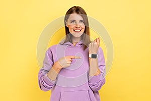 Woman with brown hair pointing finger on watch on her wrist looking at camera with toothy smile.