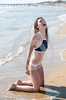 Woman bruenette is kneeling on the beach facing the sea and smiling.