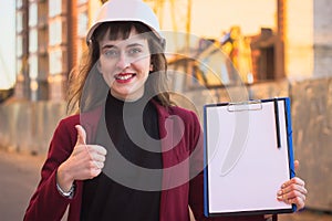 Woman builder holding blueprints, clipboard. Smiling architect girl in helmet at building background