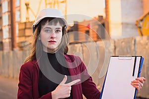 Woman builder holding blueprints, clipboard. Smiling architect girl in helmet at building background