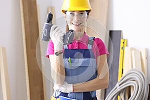 Woman builder in overalls and protective helmet holding drill in hands in workshop