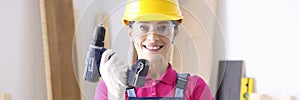 Woman builder in overalls and protective helmet holding drill in hands in workshop