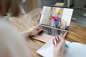 Woman builder in protective helmet waving hand from laptop screen to client