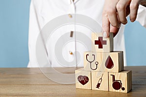 Woman building pyramid of cubes with different icons on wooden table against light blue background, closeup. Insurance concept