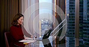 A woman in a Burgundy dress sits at a round table near a large window. she communicates through a video link on a laptop